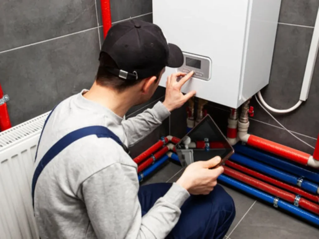 Techncian in black cap and overalls using tablet and hand to program a white wall-mounted tankless water heater with visible red and blue pipes.
